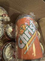 Close-up photo of a single orange soda can style money bank wrapped in clear plastic. Front branding clearly visible reading 'Christia' with slogan 'quenching thirst for your soul'.
