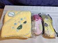 Photo showing a yellow hooded baby towel with a turtle and starfish embroidery alongside two pairs of new kids' slippers still in packaging — one pink with a strawberry design and one yellow with a sun design.