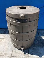 Brown plastic rain barrel in outdoor setting, showing top lid with circular opening and spigot at base.