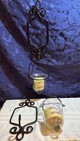 Photo showing one wrought iron wall hanger hanging with glass candle holder and vanilla scented candle, plus a second hanger and candle holder with candle on table.
