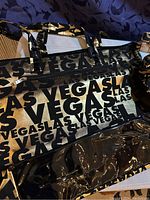 Close-up photo showing a large glossy duffel bag with a repeating 'Las Vegas' print in black and gold with shiny black patent trim.