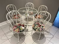 Overview of five white painted steel chairs with floral upholstered seats and part of a matching round glass table.
