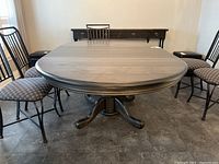 Wood composite oval dining table with pedestal base viewed from the front, showing the table top, metal frame chairs around it, and a background console table.