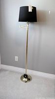 Full view of the brass colored floor lamp with brass base, adjustable arm, and black lampshade against a gray wall.