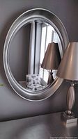 Front view of oval mirror hung on a grey wall, reflecting a chair, window shutters, and part of a table lamp with a chrome base.