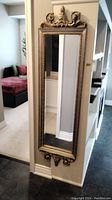 Full view of the tall narrow mirror mounted on wall, showing beveled glass and full frame with ornate top and bottom.