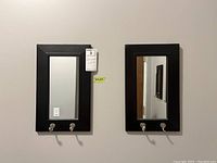 Front view of the two black framed rectangular mirrors, each with two metal hooks below the mirror surface. Both mirrors are mounted on a light grey wall, one shows a broken hook.