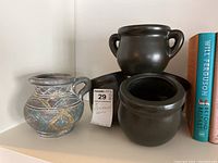 Four stoneware items: one decorated pitcher with geometric patterns and three dark brown vases of varying sizes. Shelved beside books for size reference.