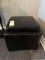 Front view of black vinyl upholstered storage stool with a cushioned removable seat.