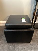 Front view of black vinyl upholstered storage stool with padded seat featuring clean straight stitching and smooth finish.