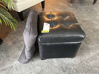 Side view of the black vinyl storage stool with button-tufted top and spare dark cushion leaning against it. Shows wear and peeling on vinyl edges.