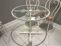 Round painted steel dining table with removable glass top, showing top with glass in place and 2 floral cushioned dining chairs in white metal frames.
