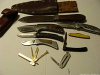 Seven folding knives and cutters arranged side by side, with two leather sheaths above, displaying various handle materials including wood, bone, and metal.