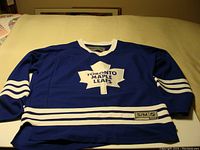 Blue Toronto Maple Leafs hockey jersey laid flat showing front logo, size tag, and striping details.