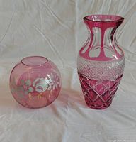 Both cranberry vases shown side by side on white cloth background. Left is round cranberry flash rose bowl with floral hand-painted design, right is tall Bohemian cut crystal cranberry vase with diamond pattern.