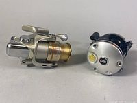 Side view of Daiwa Emblem-X 2000C spinning reel and Abu Garcia Ambassadeur 5500-C3 baitcasting reel placed side by side on white background.