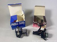 Two fishing baitcasting reels with their original boxes; one blue-gray, one black with red and gold accents.