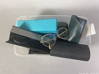 Plastic bin with various eyeglass cases and one pair of gold-tone glasses on top