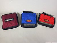 Three Tackle Logic branded fishing tackle bags in burgundy, blue, and red, zippered with mesh panels shown from the front.