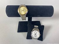 Two men's wristwatches displayed on black watch holders, Citizen with gold dial and Casio with white dial and two subdials