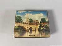Closed metal toffee tin with colorful scenic depiction of a stone bridge over a river and old buildings, labeled 'Aylesford'. Tin shows light wear.