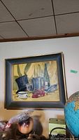 Framed acrylic still life painting hanging on a wall above various objects including a globe and a ceramic pot.