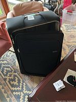 Front view of large black suitcase with zippered front pocket and handle on top, placed on patterned rug near furniture.