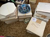 Stack of white and cardboard boxes containing collectible plates, one plate visible on top depicting a warrior on horseback at night with a spear
