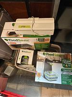Photo showing two FoodSaver vacuum sealer machines in original boxes, handheld vacuum sealer and white vacuum sealing machine without box.