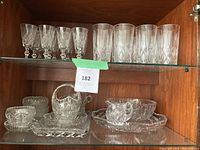 Two shelves of assorted crystal items including goblets, large glasses, bowls, and serving pieces arranged inside a wooden cabinet.