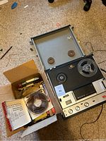 Photo showing vintage Sony reel-to-reel tape recorder placed on carpet floor next to a box containing multiple tape reels and instruction manuals.
