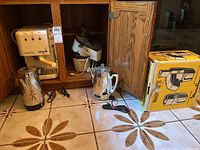 Wide shot of all appliances in and beside wooden cabinet including the Cuisinart coffee machine, Osterizer food processor, two stainless steel electric percolator kettles, and packaging box for a vintage hand mixer.