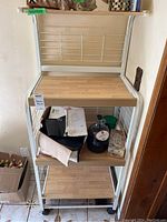 Front view of bakers rack showing four MDF shelves with items on some shelves for scale, metal frame painted white with wire side and back panels, caster wheels visible at bottom.