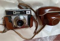 Camera front view showing Agfa Color Apotar 1:2.8/45 lens with lens glass reflecting light, the camera body with leather half-case attached, and the Agfa logo visible on the front of the camera.