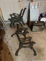 Four cast iron bench legs displayed side-by-side on a garage floor showing ornate scrollwork and curved armrests.