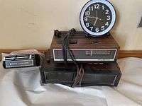 Three items displayed including the Lloyds flip-digit clock radio, GE electronic clock radio, and Sanyo underdash eight track player with mount, all placed on a white surface against a beige wall.