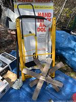 Yellow hand truck dolley in original box with two wooden bases on wheels in backyard setting on blue tarp.