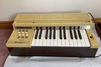 Front and side view showing 25-key keyboard, 6 major chord buttons (A, B♭, B, C, D, F), volume slider, and power switch on the top right.