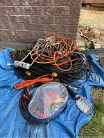 Photo showing multiple extension cords including black and orange ones, jumper cables and handheld work light on blue tarp outside near brick wall.