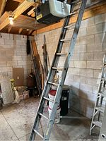 Metal extension ladder standing in a workshop with concrete block wall and wooden ceiling beams. Ladder shows some scuff marks and use wear.