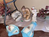 Wide shot of two blue floral wall sconces and three porcelain figurines including two kneeling females and one angel figure holding a candle.