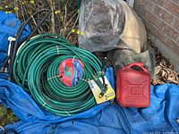 Lot contents showing multiple garden hoses and gas can on blue tarp with background brick wall and leaves.