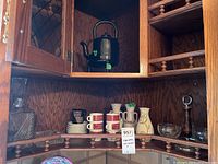 Corner cabinet shelf displaying four white espresso mugs with red designs on saucers, two ceramic vases (one beige with floral design and one smaller), a clear glass decanter with stopper, and a round clear glass ashtray.