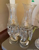 Pair of glass hurricane lamps show full view of lamp height, clear glass with hanging prisms and wrapped cords at base.