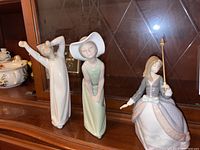 Three Lladro porcelain figurines arranged on a wooden shelf, showing different angles and poses of women in fine dresses.