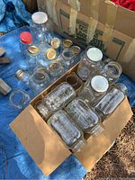 Clear glass mason jars in cardboard boxes on blue tarp, multiple jars with some metal and plastic lids visible.