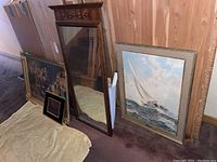 View showing large wood framed mirror with decorative top, leaning against wall, and sailboat print next to it plus other framed prints on floor.