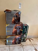 Photo showing stacked NOMA outdoor lighting boxes including carriage light and solar garden lights alongside a Laflame carousel light box on tiled floor.