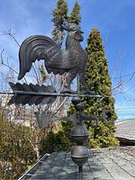 Photo of metal rooster weather vane showing detailed rooster figure and directional markers on rooftop under clear sky.