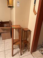 Two wooden plant stands of different heights and widths placed side by side on a tiled floor next to a wall.